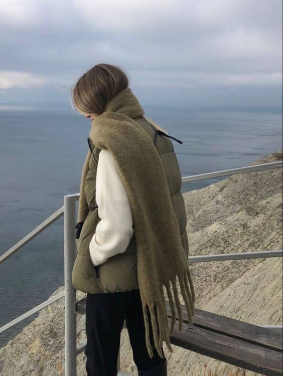 Woman in cozy winter outfit overlooking ocean from cliffside, wrapped in warm scarf and puffer vest.