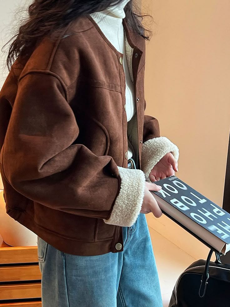 Woman in a brown shearling jacket holding a book, wearing a white turtleneck and blue jeans indoors.