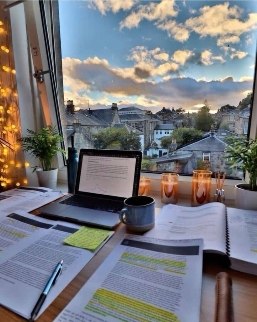 Cozy study setup with a laptop, papers, coffee, and a scenic window view of rooftops under a blue sky.