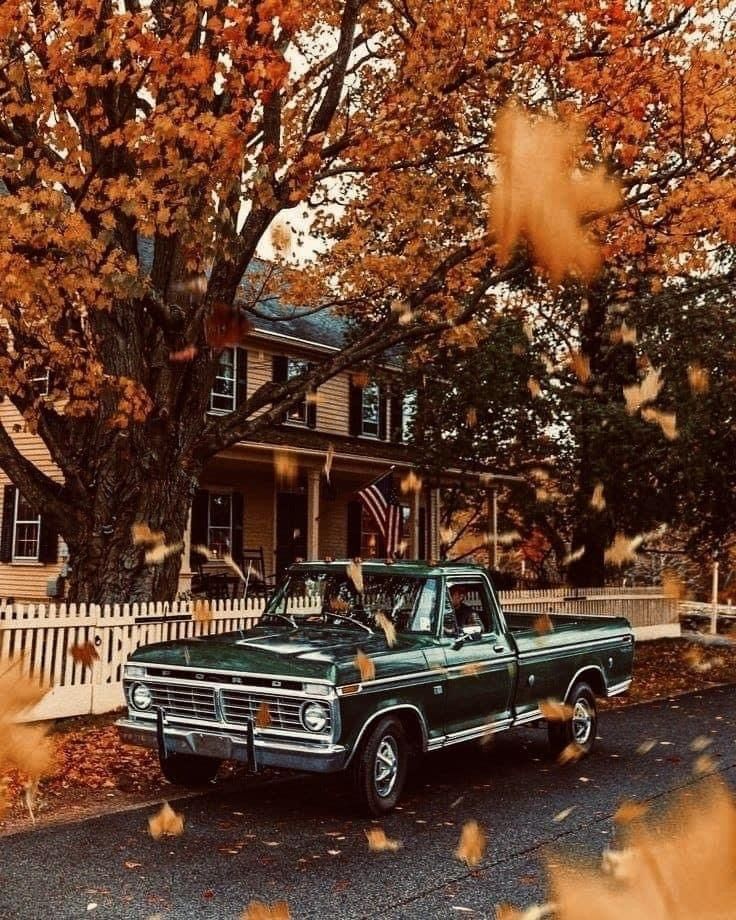 Vintage green truck under vibrant autumn leaves by a classic house with picket fence.