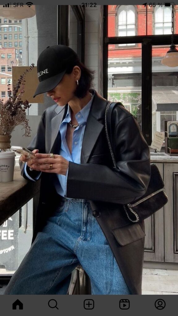 Trendy woman in a leather jacket and cap checking her phone at a stylish café window seat.