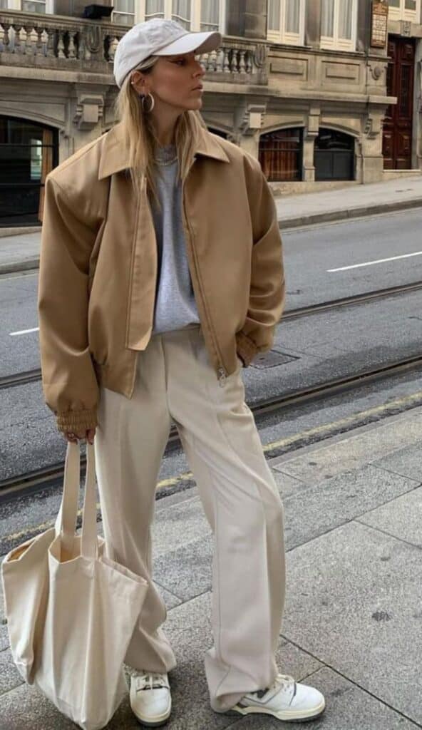 Stylish woman in oversized tan jacket, beige pants, and cap, holding a tote bag on city street. Casual urban fashion.