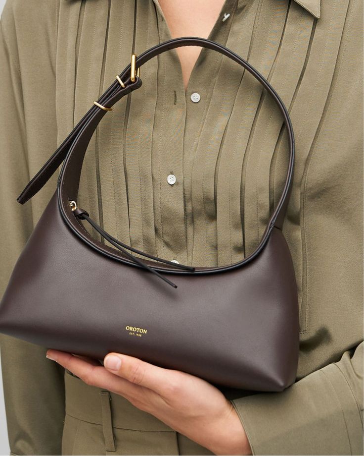 Woman holding a stylish brown leather handbag with a minimalist design against a green outfit.