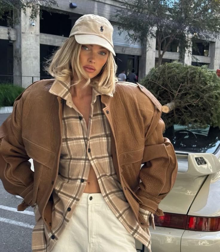 Stylish woman in plaid shirt, brown jacket, and cap, standing by sports car outdoors.