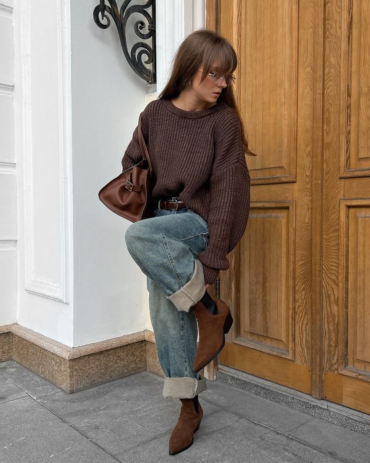 Fashionable woman in cozy sweater and jeans with brown boots outside a wooden door, stylish autumn outfit.