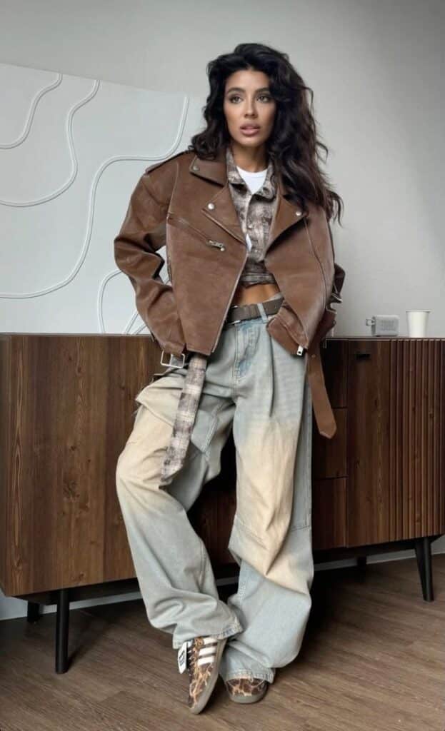 Fashionable woman in brown leather jacket and baggy jeans, standing confidently indoors.