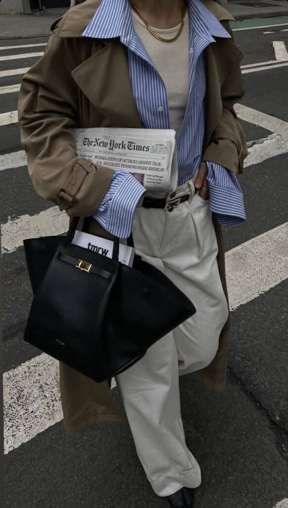 Fashionable street style: person in trench coat and striped shirt holding a newspaper and black handbag on crosswalk.