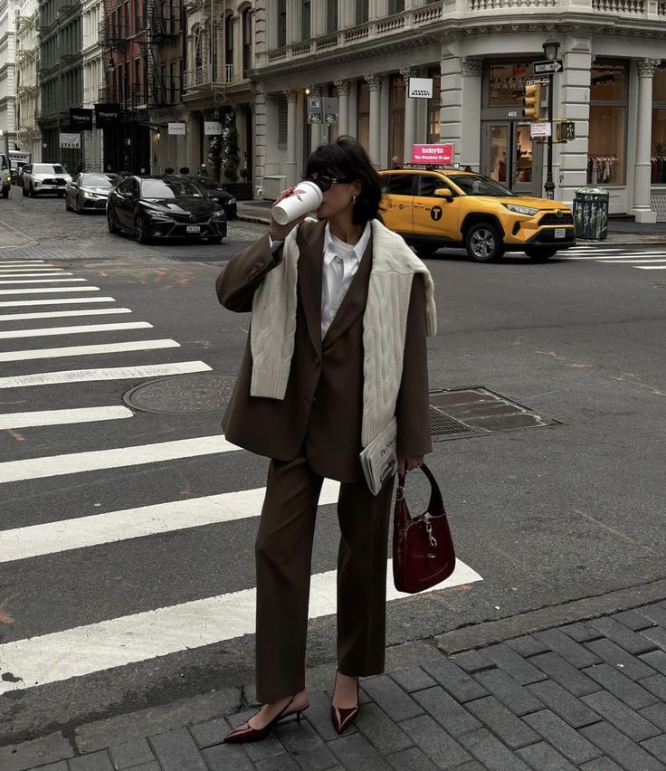 Stylish person in brown suit and heels sipping coffee on a city street crosswalk with yellow taxi in background.