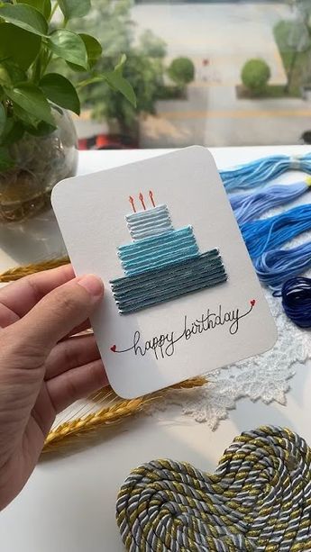 Handcrafted birthday card with a yarn cake design, held by a person, surrounded by embroidery threads.