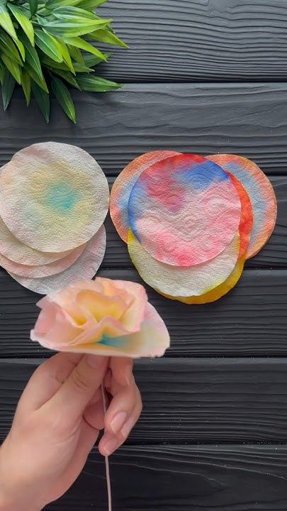 Hand holding DIY flower made from colorful painted coffee filters on a dark wooden surface.