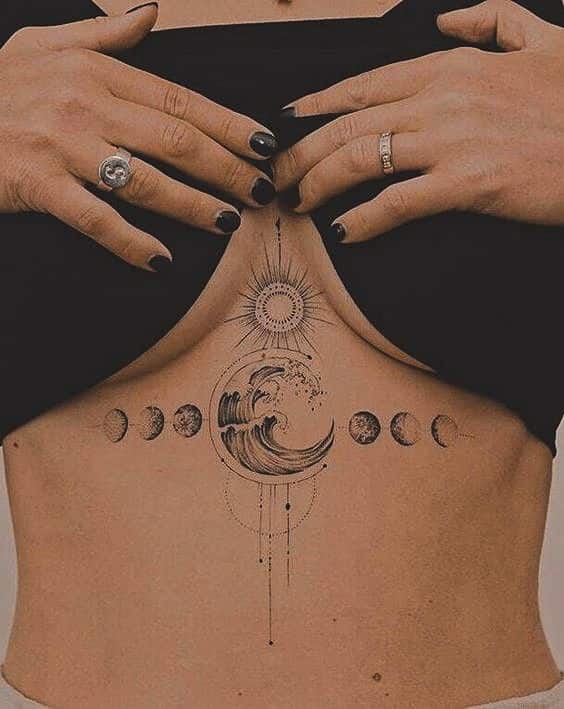 Intricate tattoo of ocean wave and lunar phases on torso, hands with black nails adjusting shirt.