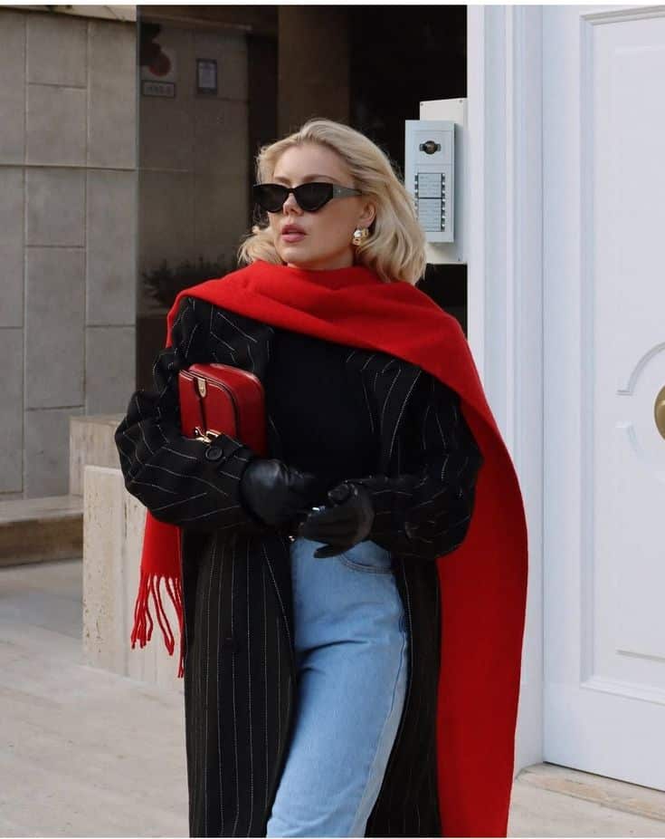 Fashionable woman in sunglasses, red scarf, and chic coat outside, holding red purse, urban style.