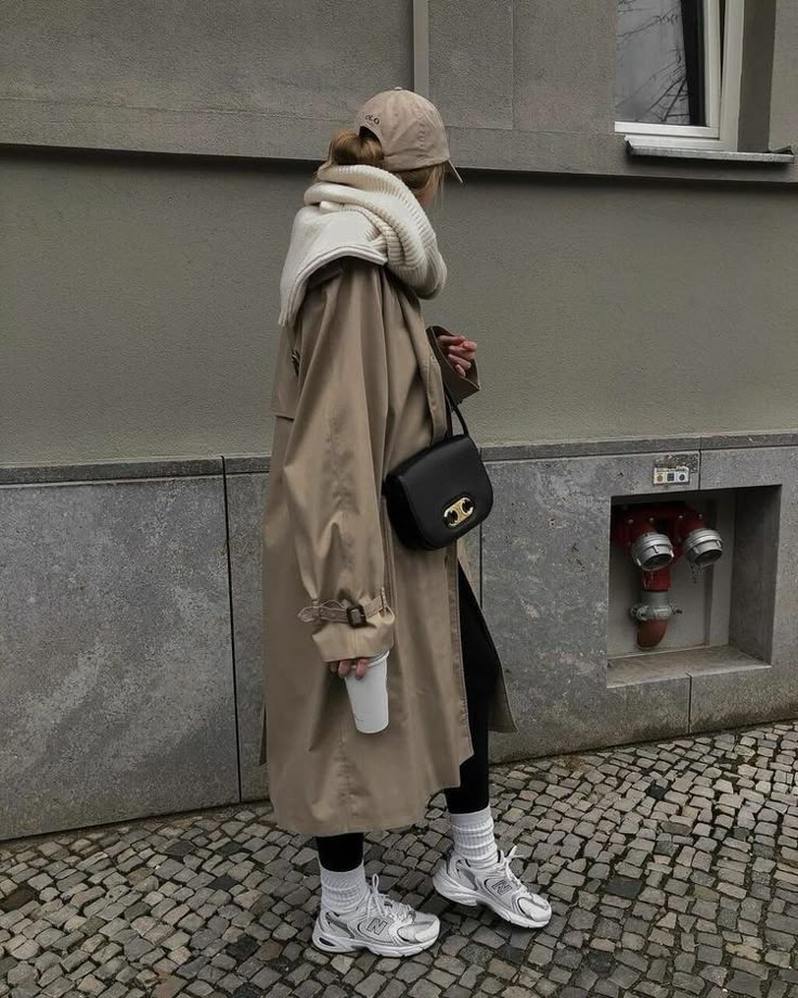 Person in casual street style with beige trench coat, scarf, cap, and sneakers, carrying a black bag and coffee cup.