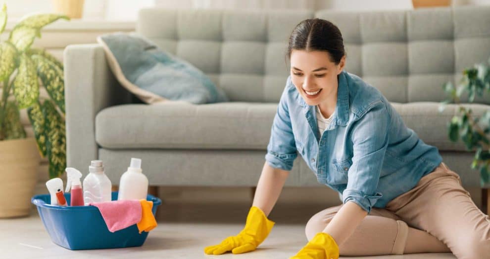 Smiling woman cleaning living room floor with gloves and supplies, enjoying home cleaning routine.