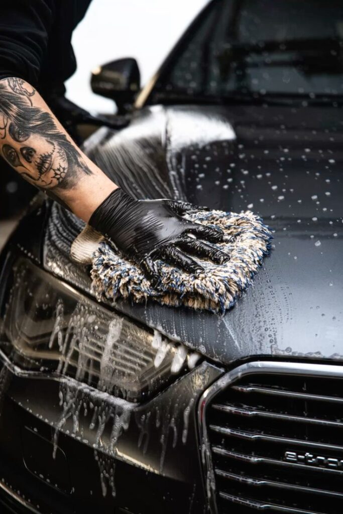 Tattooed arm in glove washing black car with foamy sponge in close-up.