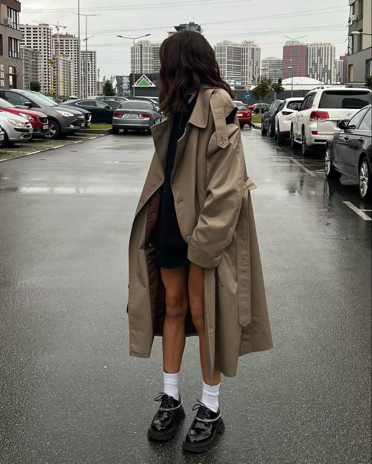 Person wearing long beige trench coat, black outfit, and loafers in a city parking lot on an overcast day.
