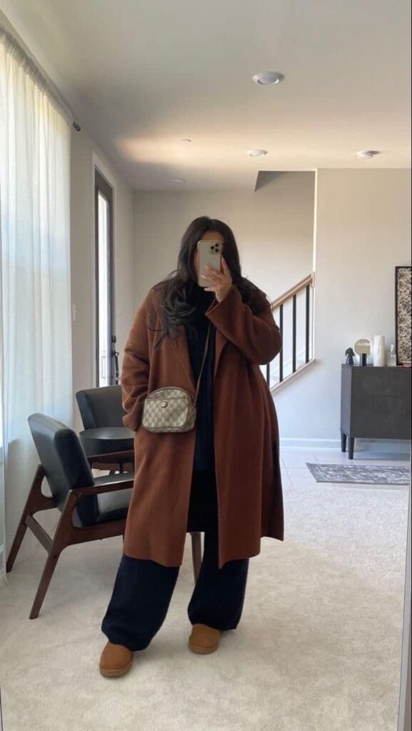 Woman in brown coat and purse taking selfie in modern living room with mirror reflection.