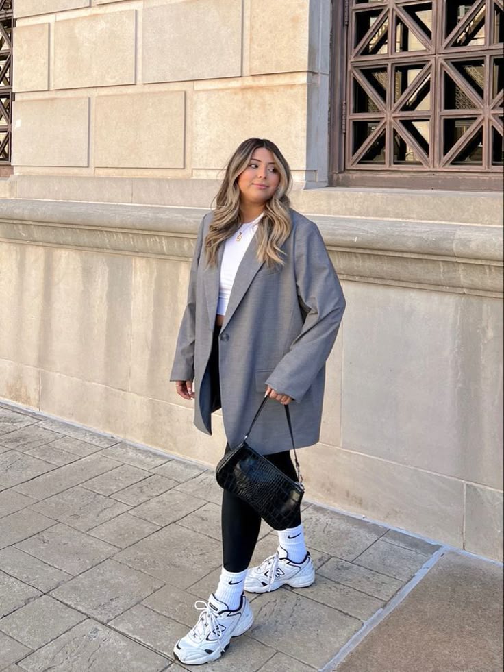 Woman in oversized blazer with sneakers, black leggings, and handbag walking by a building wall.
