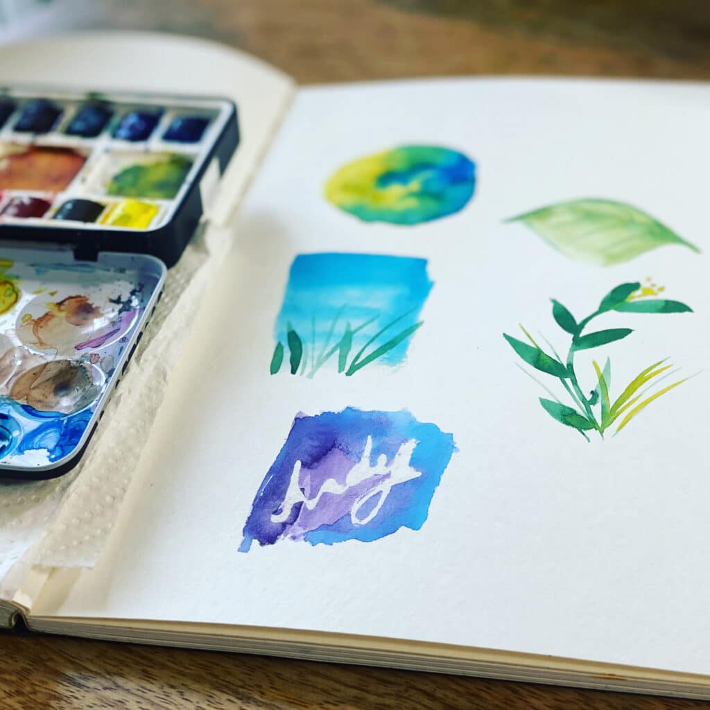 Watercolor art journal with paint swatches and a palette on a wooden table.