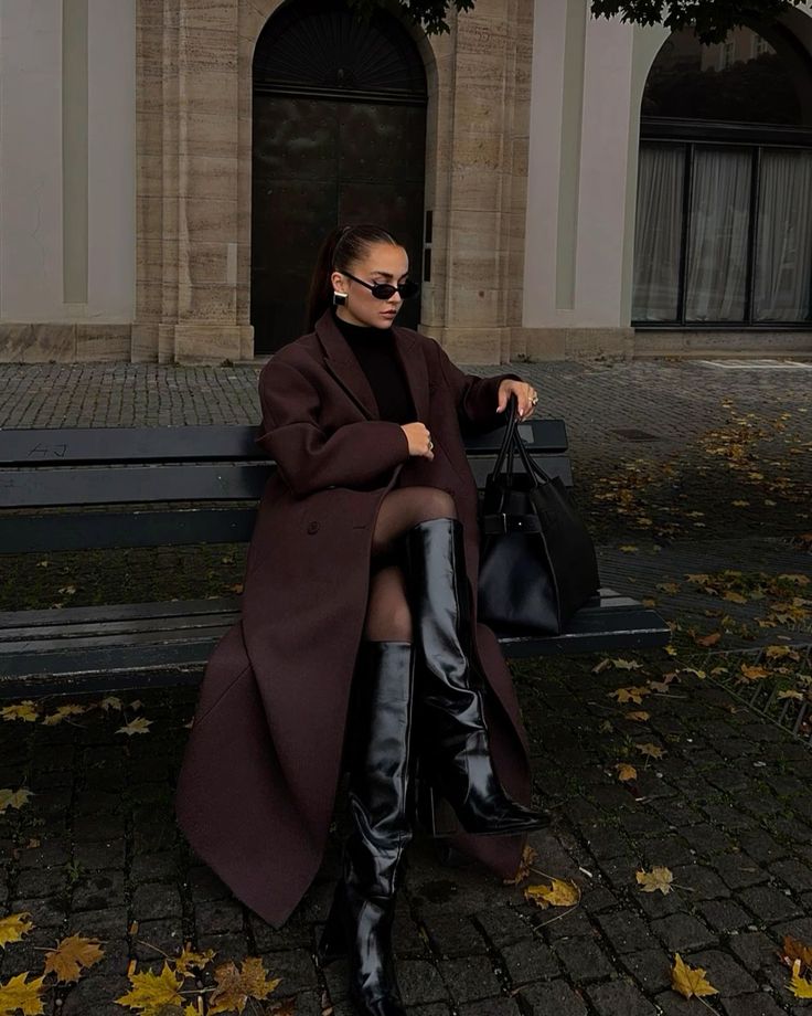 Stylish woman in dark coat and sunglasses sitting on a bench, wearing black knee-high boots, with a large handbag.