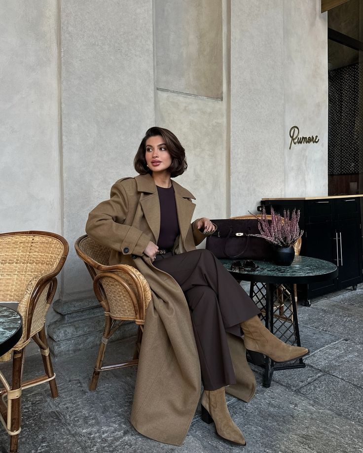 Stylish woman in a chic brown outfit and long coat sitting at an outdoor café, exuding confidence and elegance.
