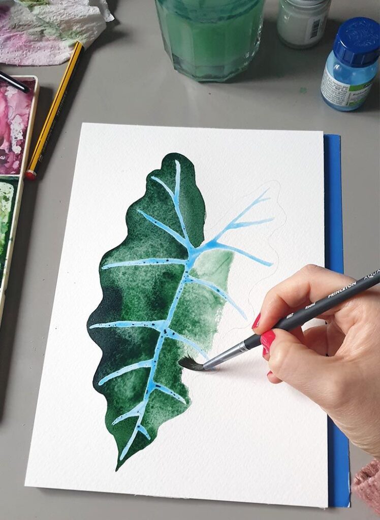 Artist painting a green leaf in watercolor on paper, with paints and brush visible nearby.