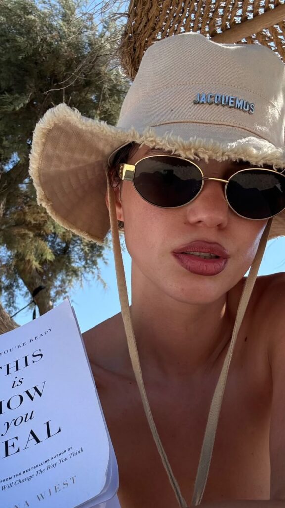 Person in sunglasses and hat holding a book outdoors on a sunny day, enjoying summer vibes.