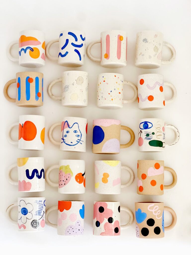 A collection of 20 colorful, uniquely painted mugs arranged in four rows on a white background.