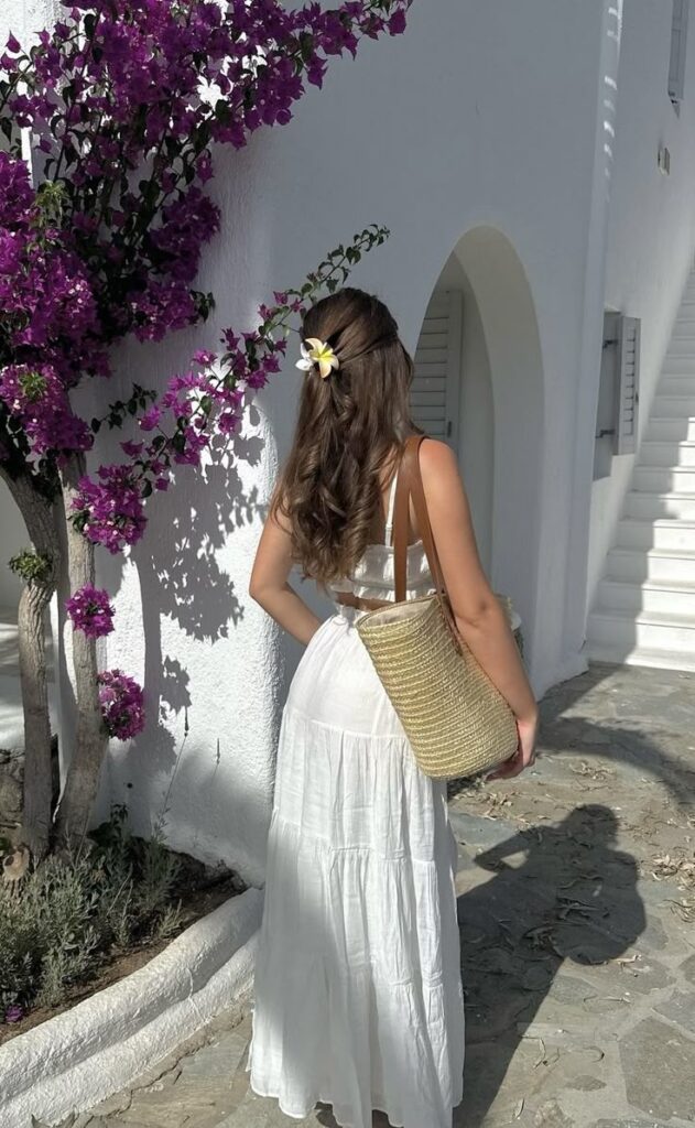 Woman in white dress with a flower in hair, standing by bougainvillea on a sunny day.