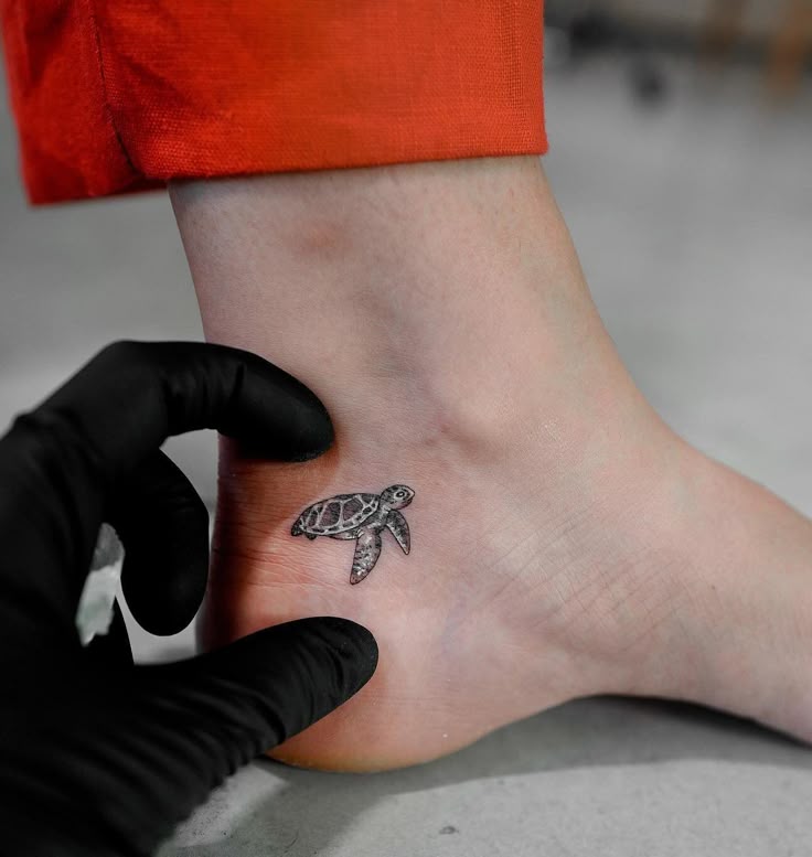 Close-up of a small sea turtle ankle tattoo being touched up by an artist in black gloves, red pants visible.