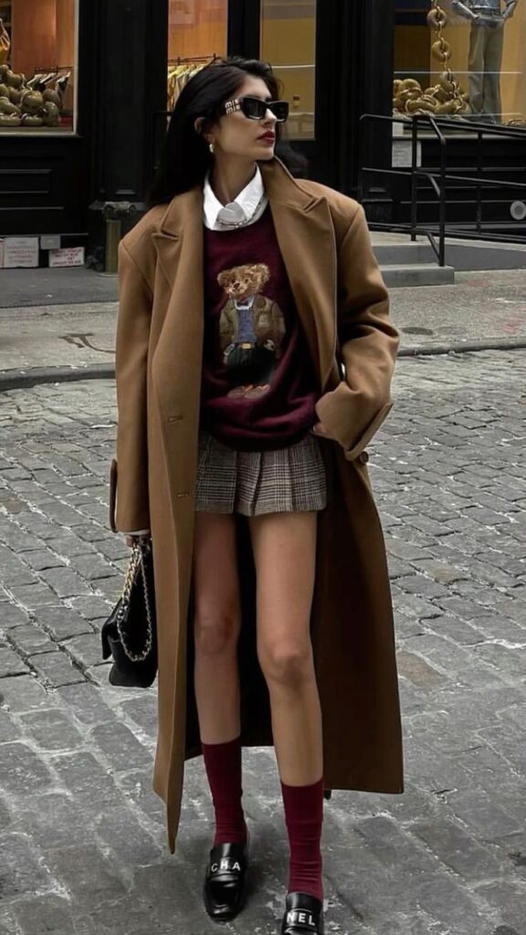 Fashionable woman in street style outfit with long coat, bear sweater, plaid skirt, and sunglasses.