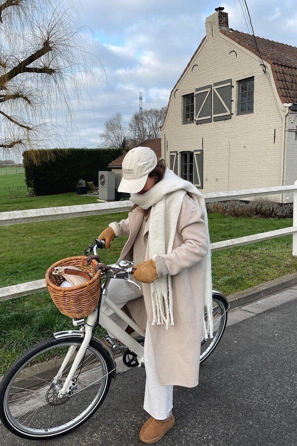 Person in winter attire biking near a rustic house, wicker basket on front of bike.