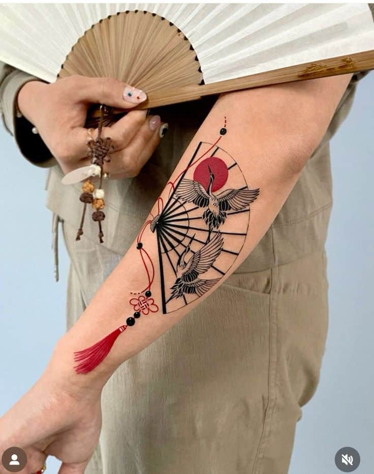 Tattoo of crane and red sun design on forearm, person holding a folded fan, wearing beige outfit.