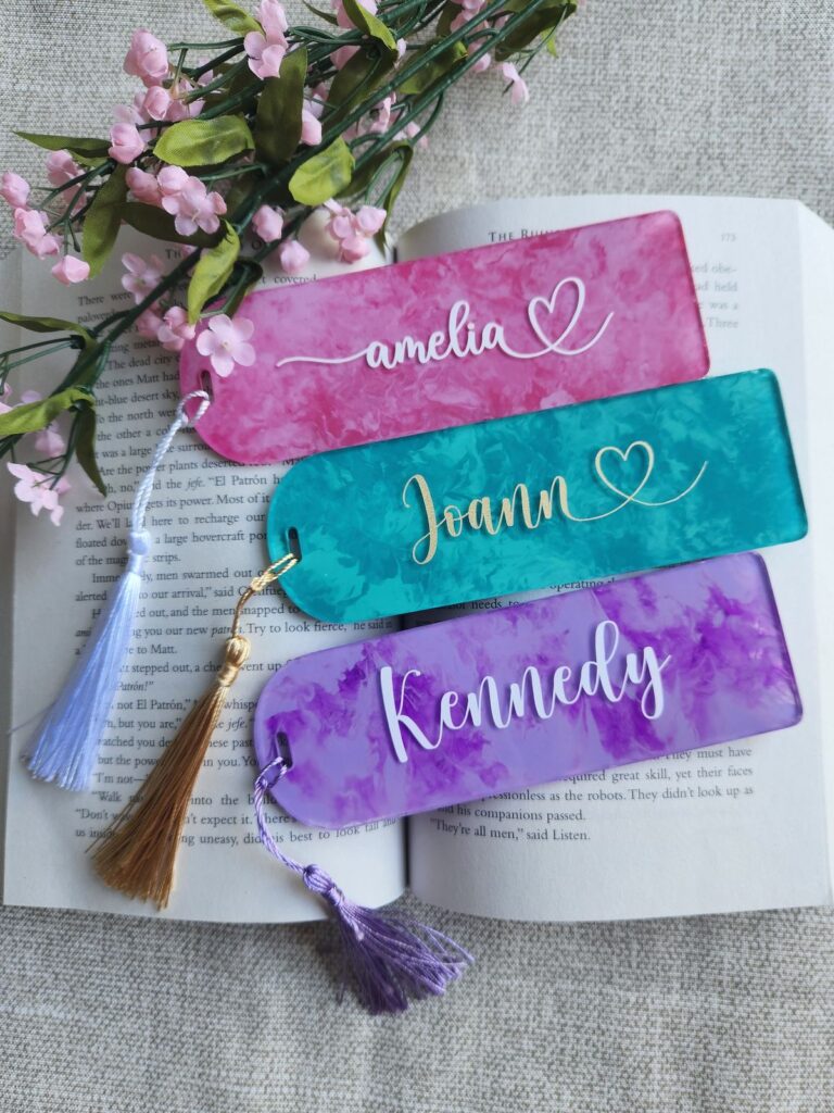 Colorful personalized bookmarks with tassels laid on an open book, adorned with pink flowers.