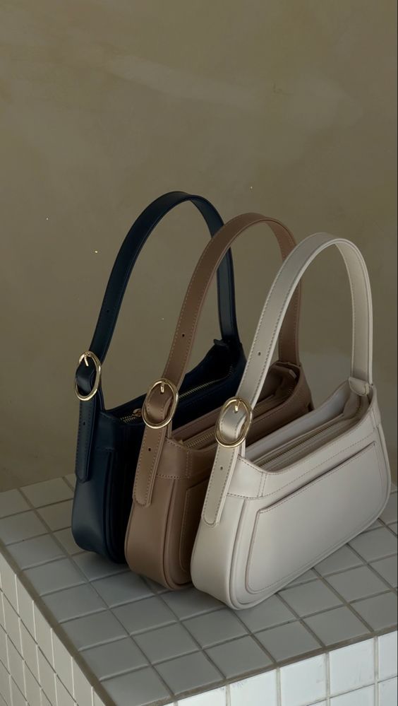 Three stylish handbags in navy, brown, and white on a tiled surface, featuring elegant buckles and modern design.