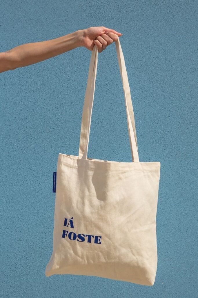 46df8770-60a0-48d6-ad3a-76fa4542997d Hand holding a beige tote bag with JÁ FOSTE text against a blue background. Eco-friendly fashion accessory. | Sky Rye Design Hand holding a beige tote bag with JÁ FOSTE text against a blue background. Eco-friendly fashion accessory.