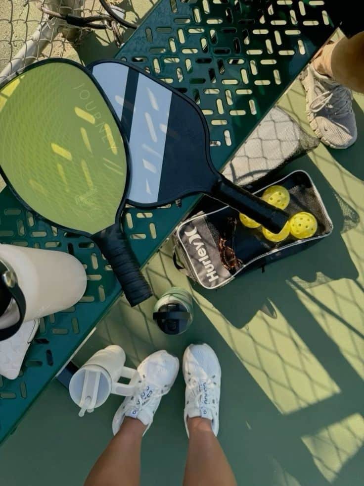Two pickleball paddles on a bench with balls and water bottles beside a person wearing white sneakers.