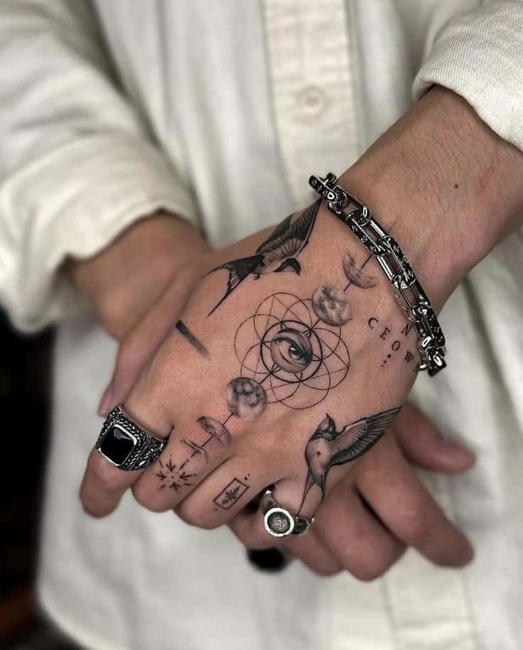 Hand with intricate tattoos featuring an eye, symbolic designs, and birds, adorned with rings and a bracelet.
