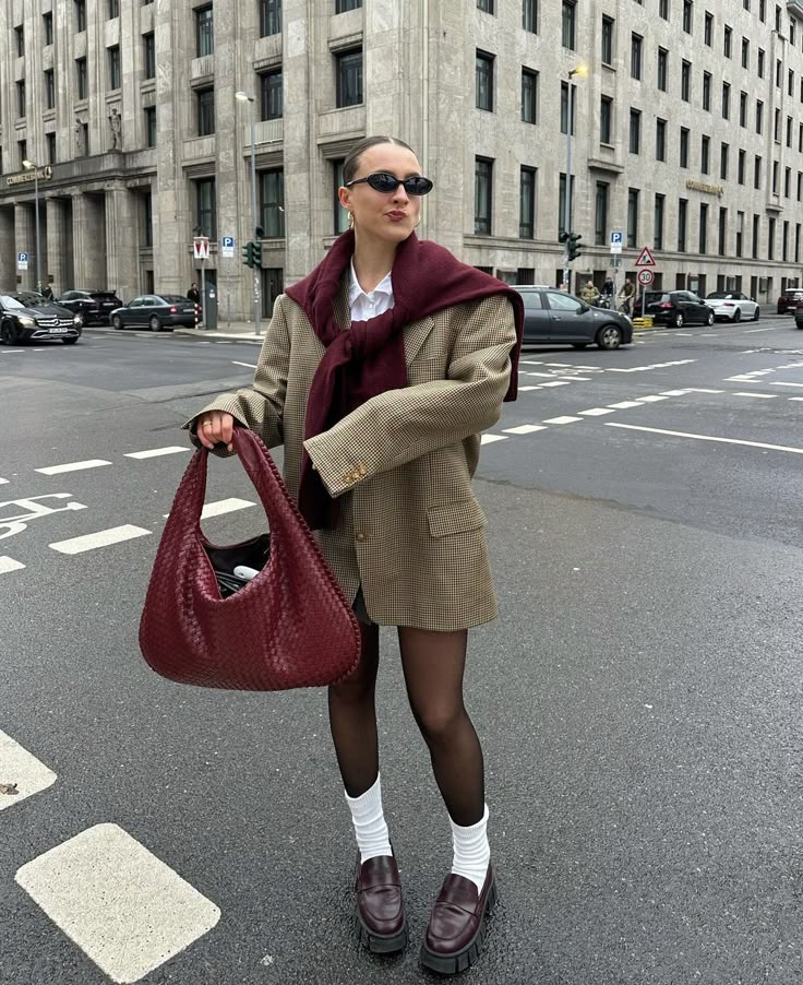 Fashionable woman in oversized blazer and loafers holding a stylish bag on a city street crossing.