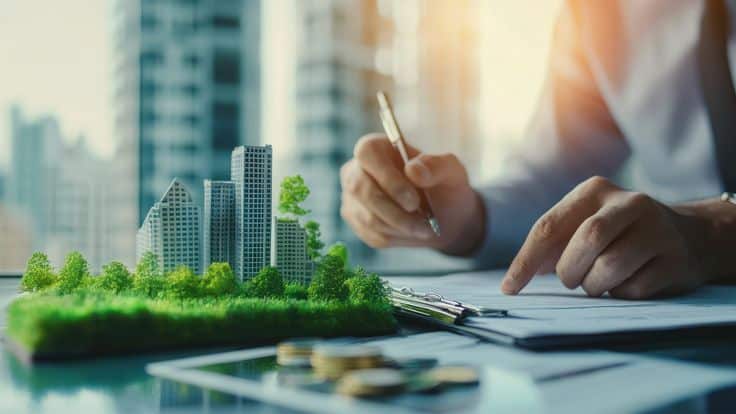 Person analyzing eco-friendly real estate model with plants on an office desk, reflecting sustainability in urban planning.
