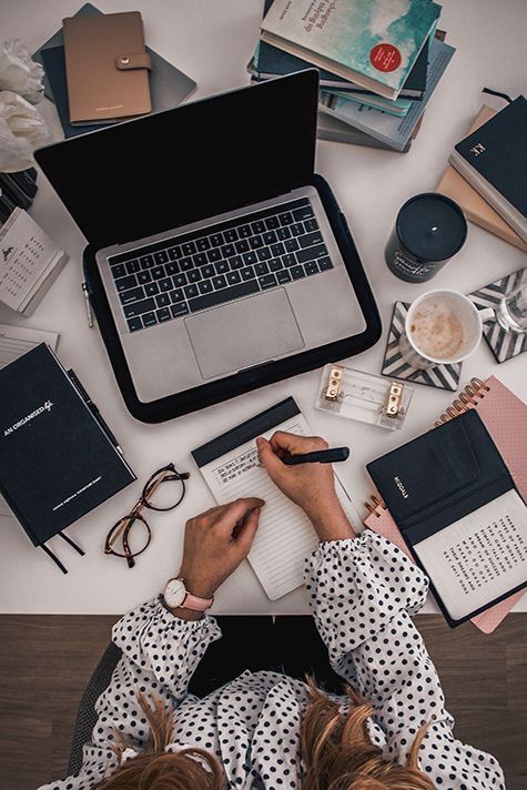 Person writing in notebook at a desk with laptop, books, glasses, and coffee cup, creating an organized workspace.