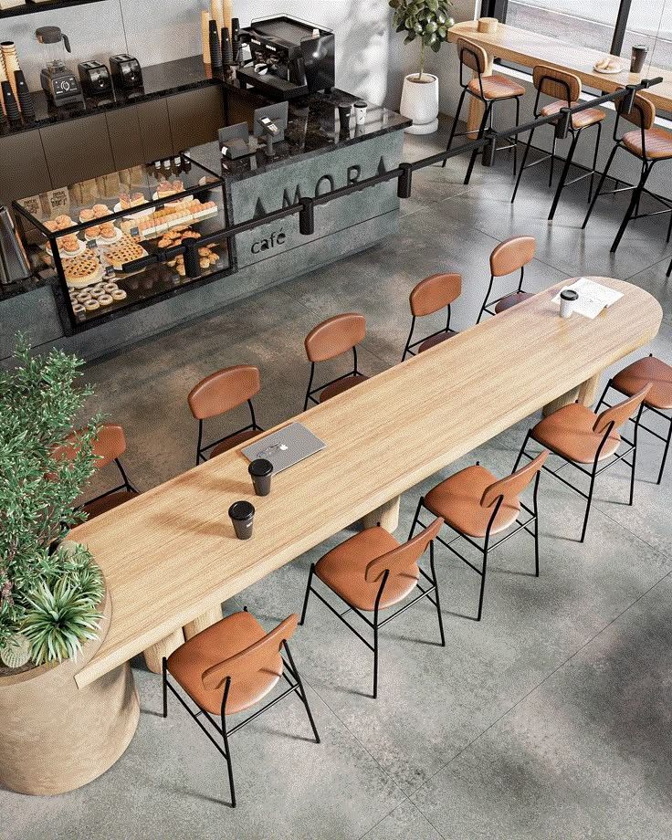 Modern café interior with long wooden table, leather chairs, and a display of pastries near the coffee counter.