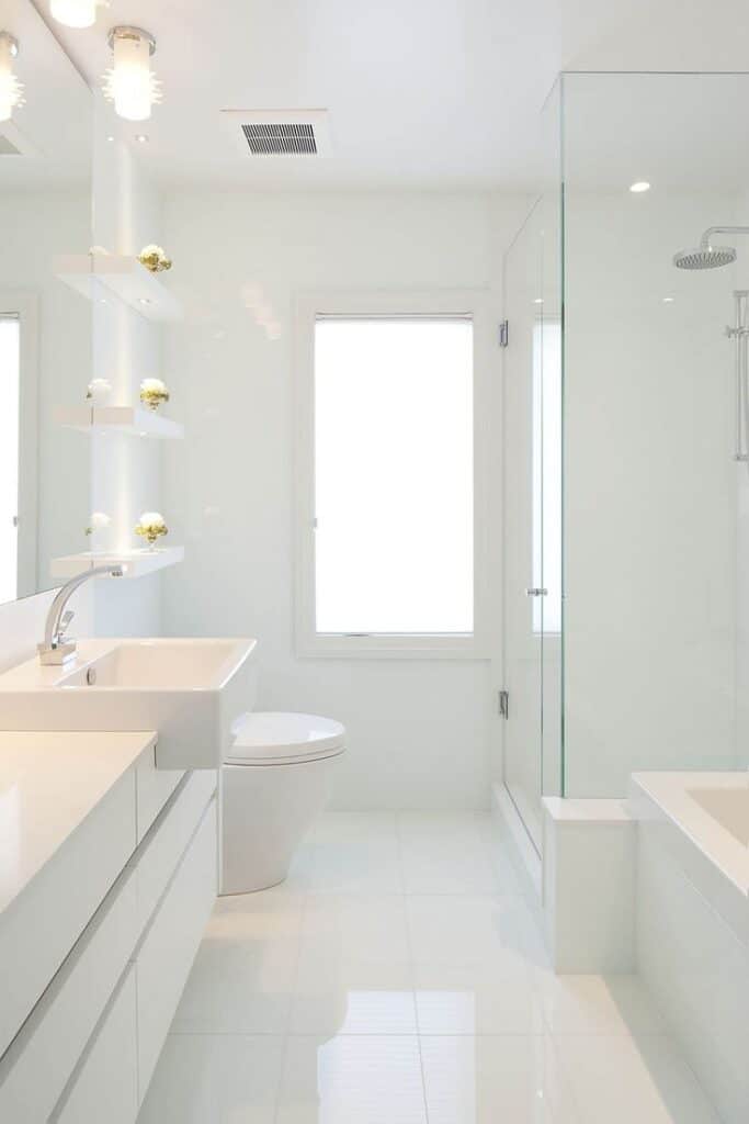 Sleek white modern bathroom with glass shower, minimalist sink, and shelves, featuring natural light from window.