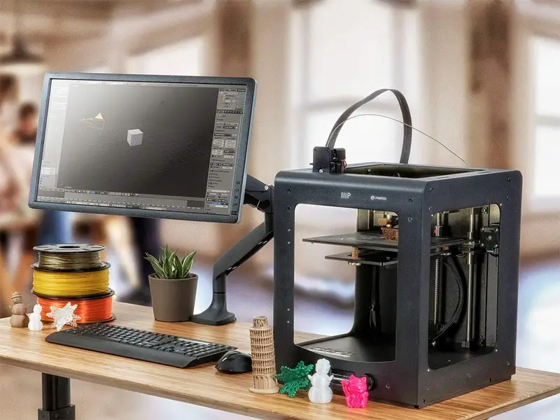 3D printer with monitor displaying design, on desk with filament spools and printed models, in modern office setting.