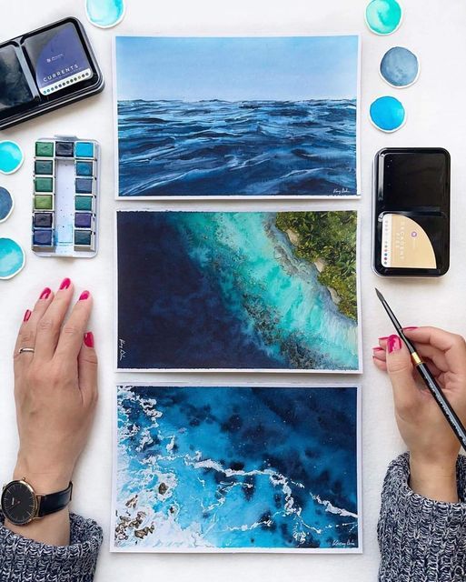 Hands with paintbrush arranging watercolor ocean paintings and paint palette on white surface.
