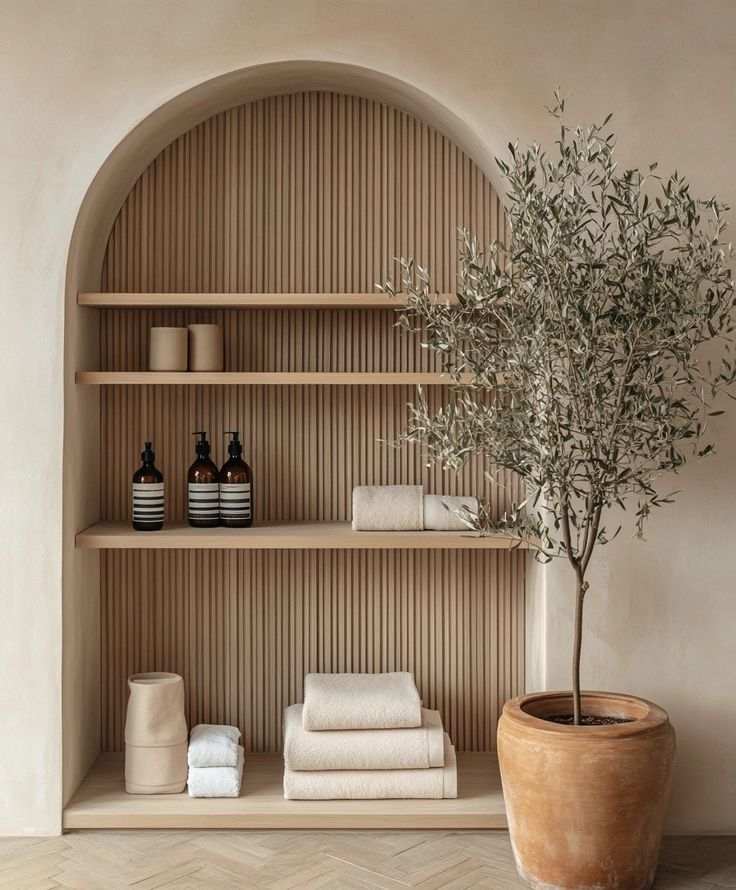 Minimalistic arch shelf with toiletries and towels, next to a potted olive tree in a serene, neutral-toned space.