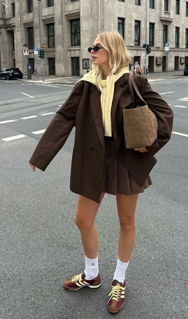 Stylish woman in a brown blazer and skirt, holding a woven handbag, standing on a city street corner.