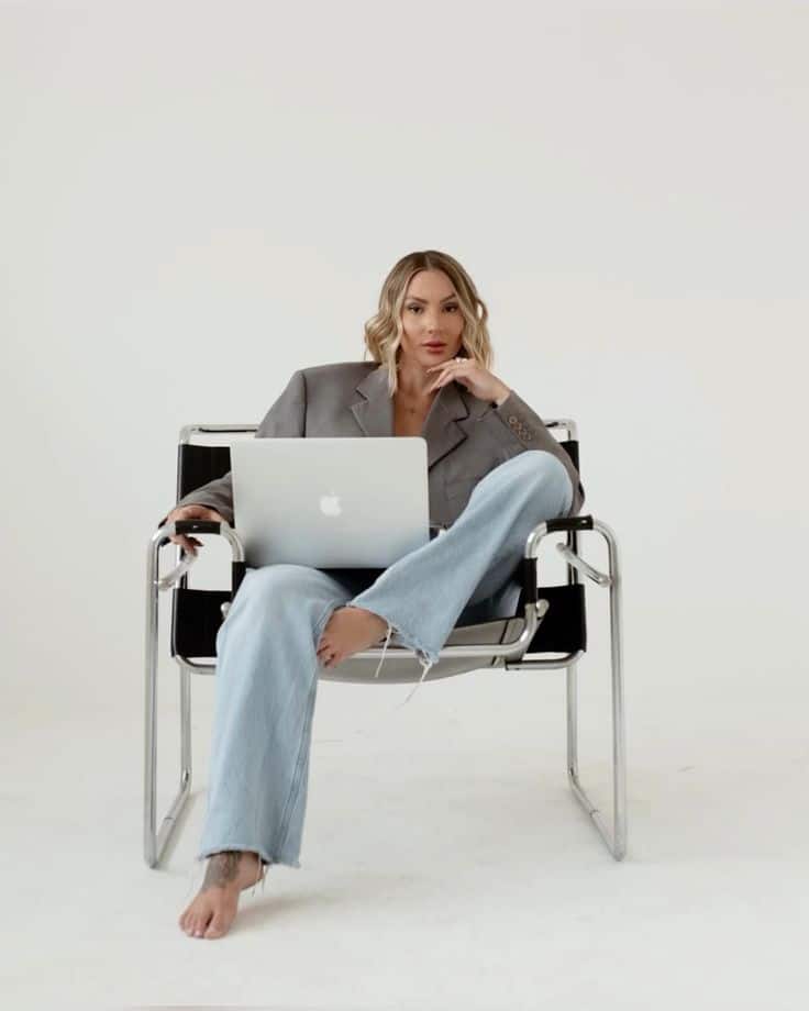 Woman in blazer and jeans sitting in a modern chair with a laptop on her lap, exuding confidence.