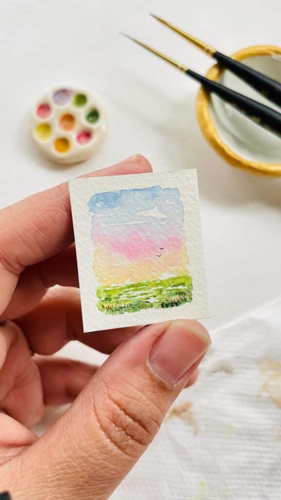 Close-up of a hand holding a tiny watercolor landscape painting with art supplies nearby.