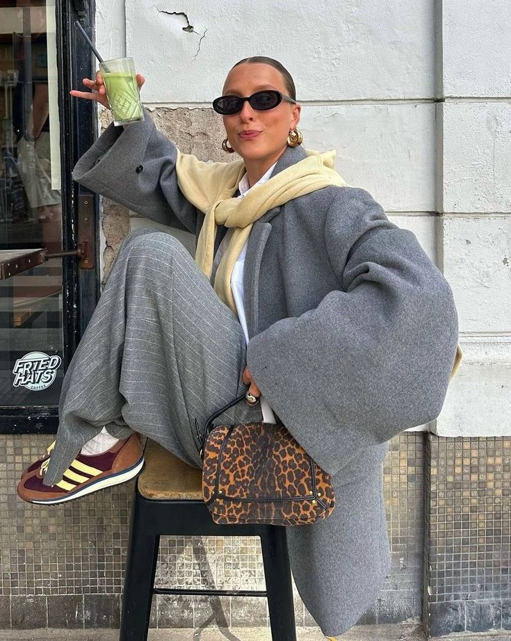 Stylish person in oversized gray coat, holding green drink, with sunglasses and leopard print bag. Urban fashion scene.